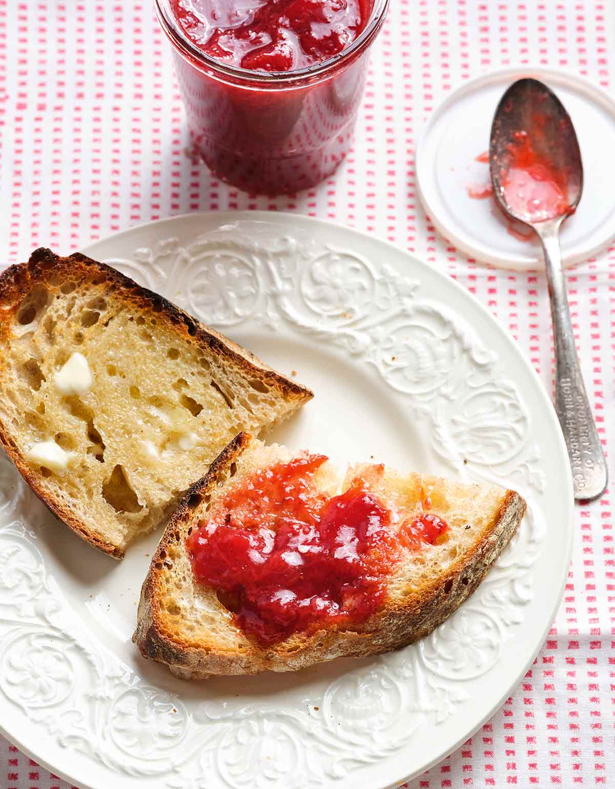 Two pieces of buttered toast, one with strawberry jam, on a white plate with a spoon and jar of jam in the background.